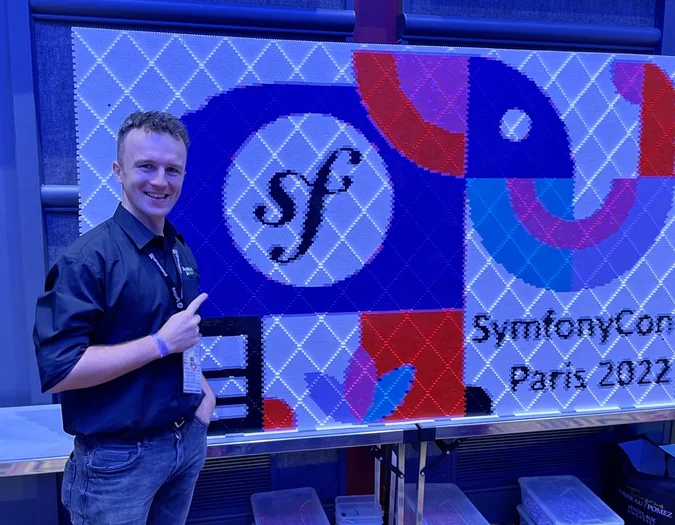Man posing next to a SymfonyCon Paris 2022 pixel art wall