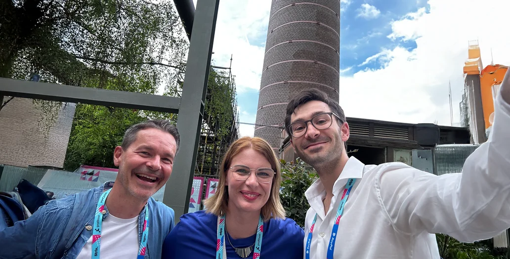 Three colleagues smiling for a selfie outdoors in front of a tall brick chimney during an event