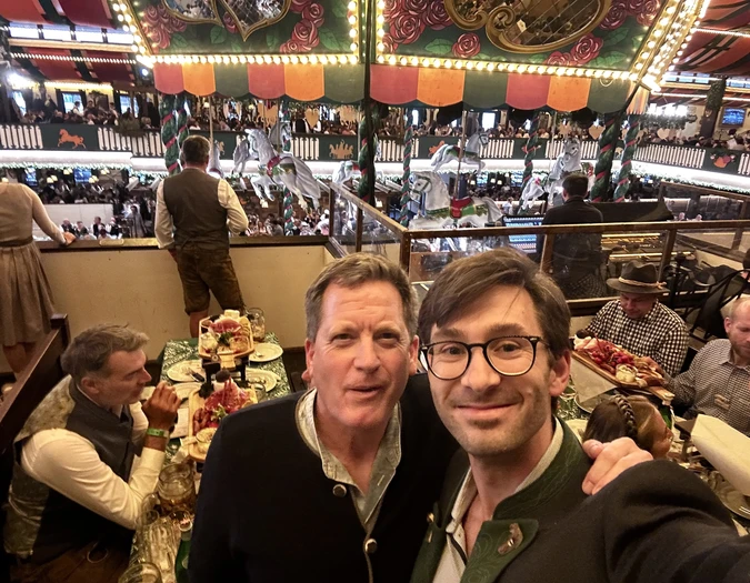 Two men posing for a selfie in an Oktoberfest tent with a carousel and traditional decorations in the background