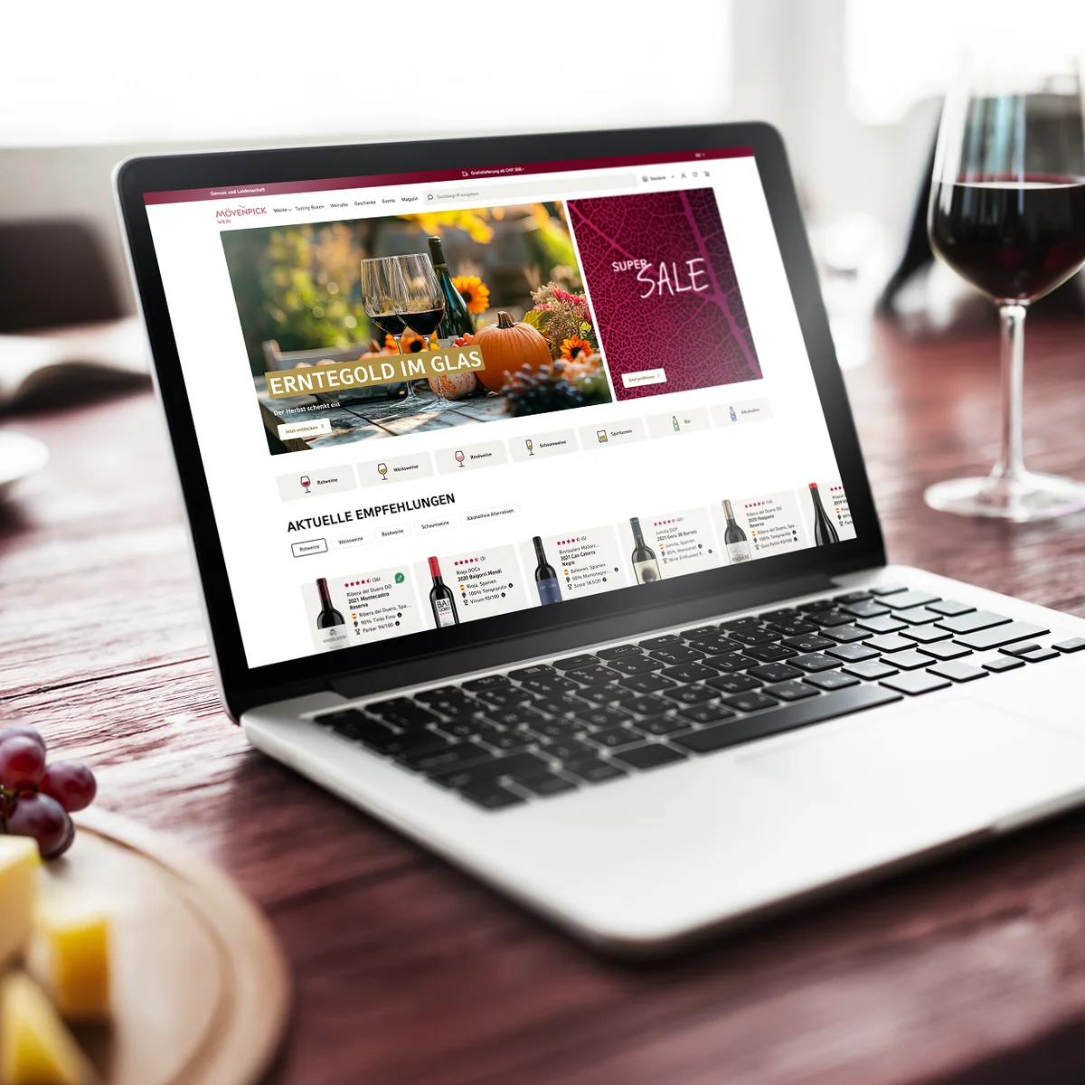Mövenpick Wein e-commerce website preview on a laptop showcasing seasonal autumn promotions and current wine recommendations, set against a cozy restaurant or dining background with a glass of red wine, grapes, and cheese