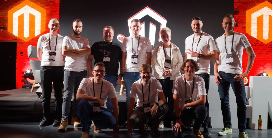 A group of team members, including CEO Jaromir, posing on a stage at Meet Magento PL Poznań with a large illuminated logo background