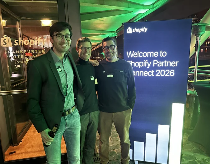 Three men posing at the entrance of a Shopify event next to a blue "Welcome to Shopify Partner Connect 2026" sign