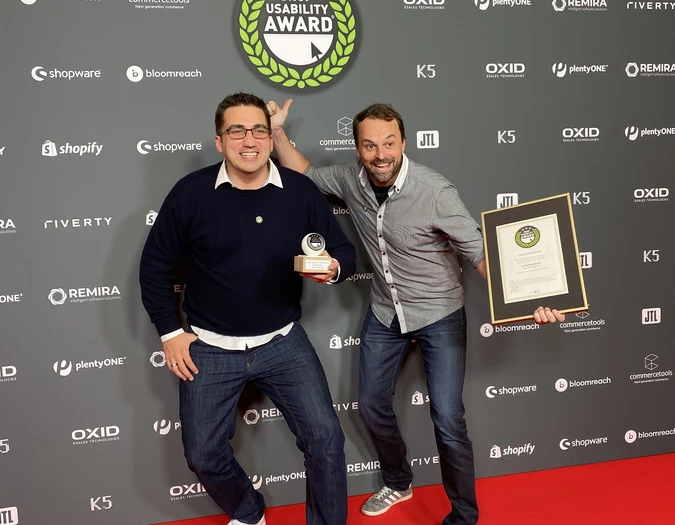 Two men posing on a red carpet with the Shop Usability Award trophy and certificate