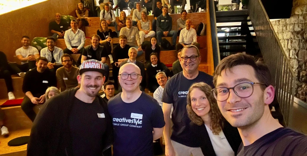 Four colleagues smiling for a selfie in the foreground with a large group of people sitting on wooden bleachers in a modern event space