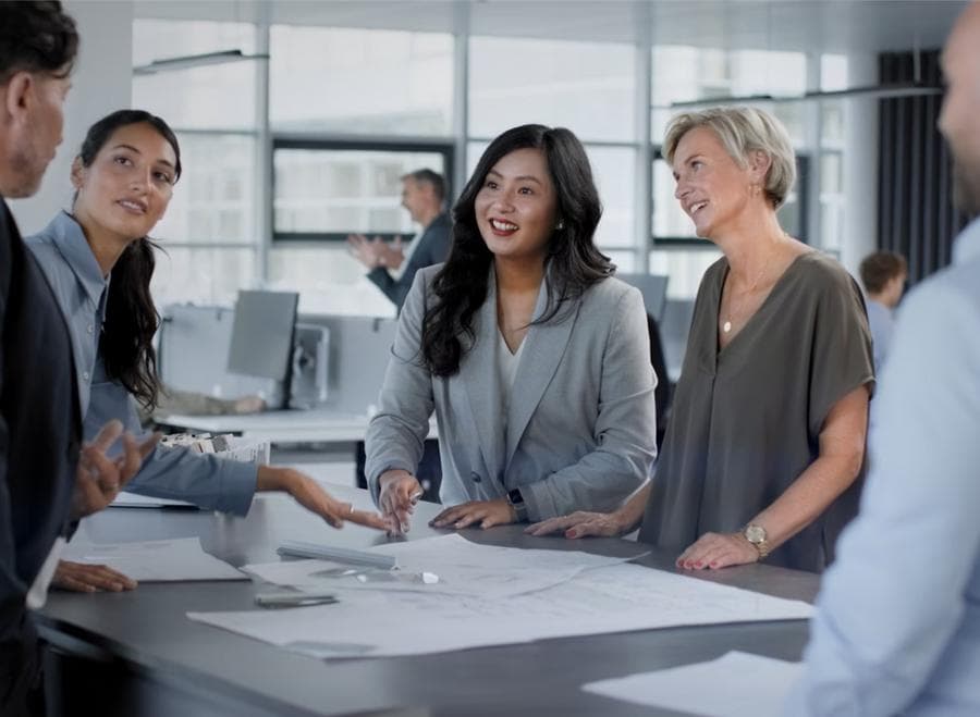 Photorgaph depicting 5 office workers: 3 women and 2 men standing around a table with blueprints, two other company employees can be seen in the background